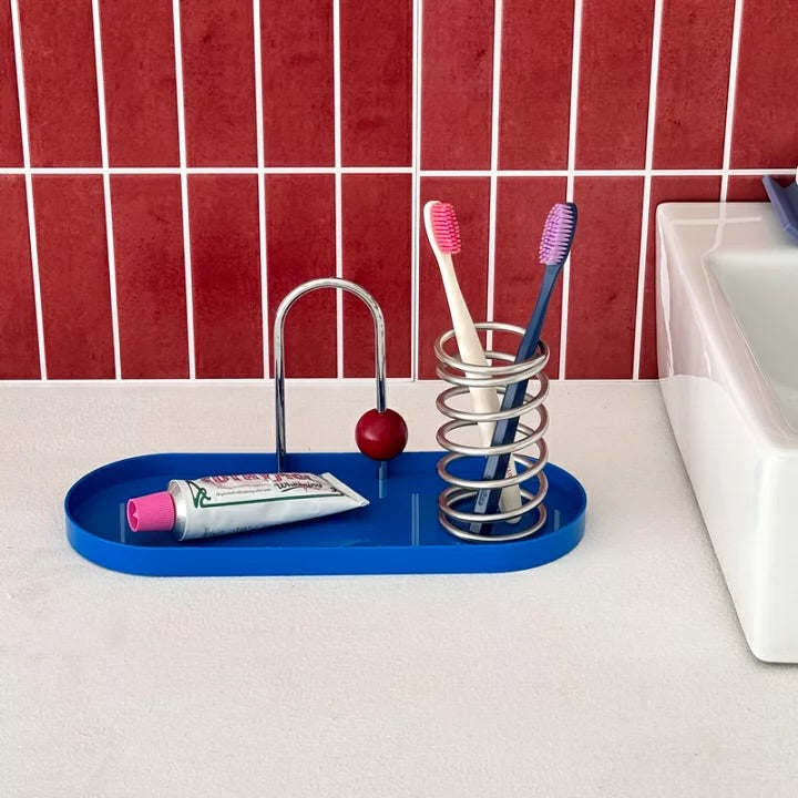 A blue bathroom organizer tray with a chrome arch holder, a spring toothbrush holder, and a small red sphere accent, holding a toothbrush, toothpaste, and two toothpicks, set on a white countertop against a red tiled wall.