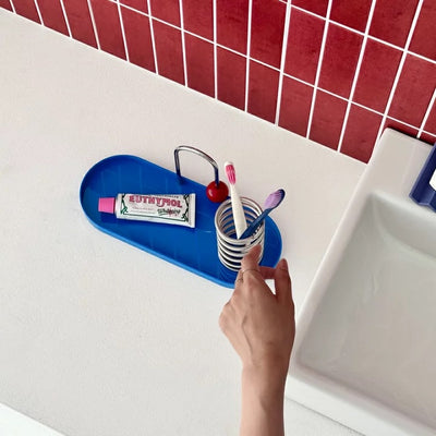 A blue bathroom toothbrush holder tray mounted on a white sink ledge, holding a toothbrush cup with two toothbrushes, a tube of toothpaste, and a small metal holder. A hand is adjusting the tray.