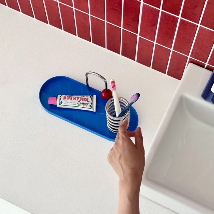 A blue bathroom toothbrush holder tray mounted on a white sink ledge, holding a toothbrush cup with two toothbrushes, a tube of toothpaste, and a small metal holder. A hand is adjusting the tray.