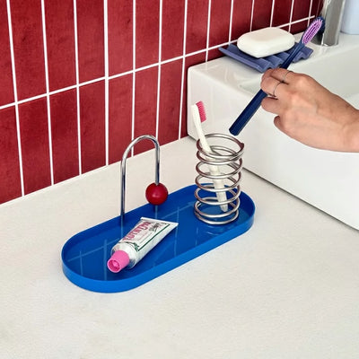 A blue bathroom toothbrush holder and organizer tray on a white countertop, holding a toothbrush in a spring cup and a tube of toothpaste on a curved stand. A hand is placing a toothbrush into the holder.
